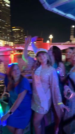 Bachelorette party on a nighttime river cruise — smiling women with a sash and captain’s hat, colorful LED party lights reflecting on the water and an illuminated city bridge and skyline in the background.
