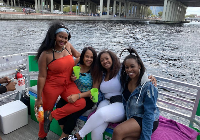 Four smiling friends on a pontoon boat along a city waterfront under a highway bridge, holding bright cups and snacks and enjoying a sunny river outing.