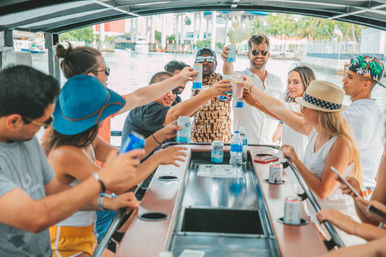 Group of friends toasting with canned drinks around a bar on a sunny party boat cruising a waterfront canal.