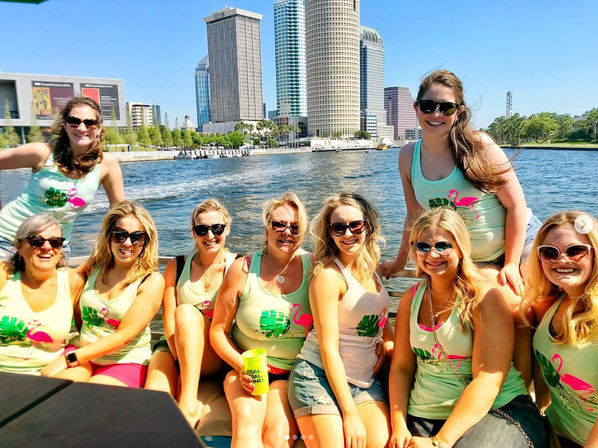 Group of women in matching flamingo tank tops and sunglasses smiling on a sunny boat ride along the Tampa waterfront with downtown skyscrapers in the background.