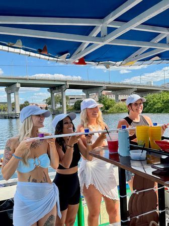 Four friends in swimsuits and white caps on a covered summer party boat holding a shot-paddle with colorful cups at a riverside bar, drinks on the counter and a concrete highway bridge over blue water in the background.