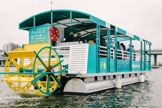 Colorful teal and yellow paddlewheel party boat with covered seating and passengers cruising an urban waterfront beneath a concrete bridge.