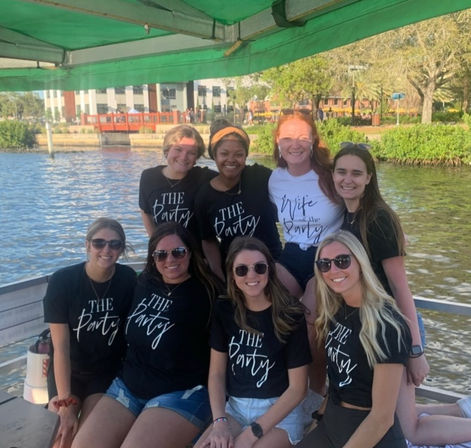 Eight friends smiling on a covered pontoon boat by a sunny waterfront, most wearing matching black "The Party" shirts and one in a white "Wife of the Party" shirt, dock and riverside buildings in the background