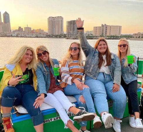 Five women in casual denim and sweaters smiling and holding drinks on a pontoon boat at sunset with a city waterfront skyline in the background.