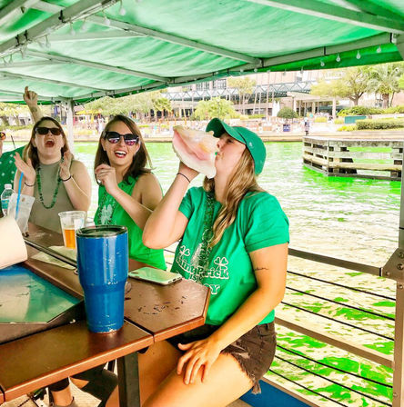 Friends in green at a covered boat bar on a waterfront river dyed bright green for St. Patrick's Day, one woman blowing a conch shell while others cheer.