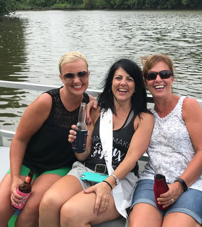 Three smiling women in sunglasses sitting close together on a boat at a lake, laughing and holding bottled drinks in casual summer clothes — carefree friends enjoying a sunny lakeside outing.