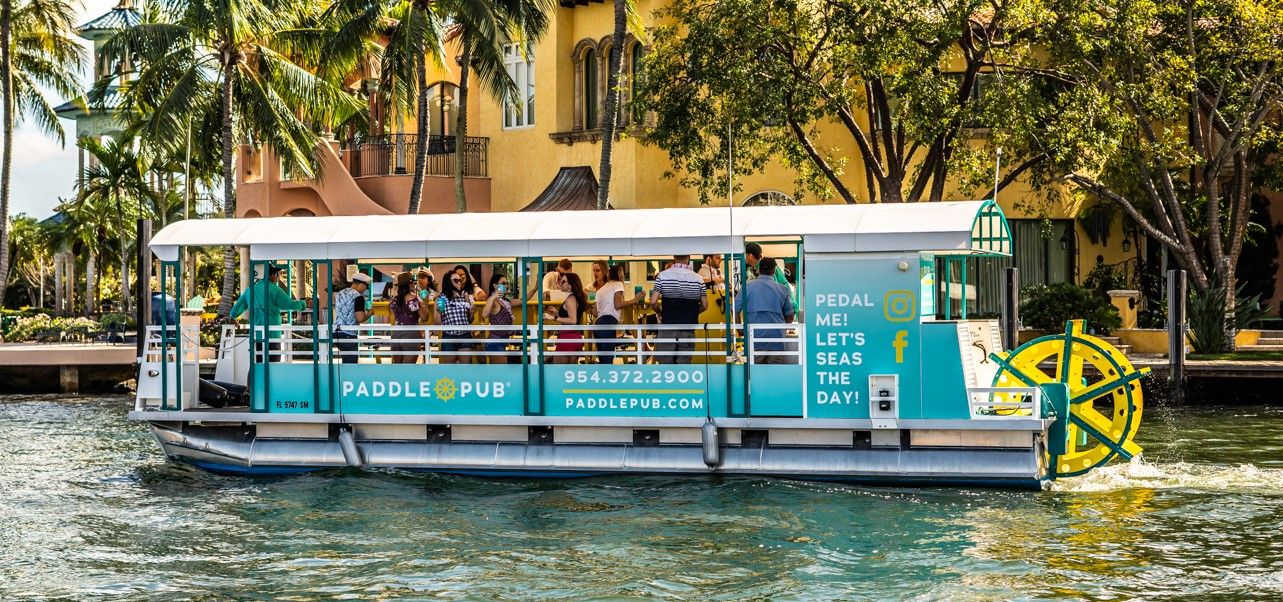 Teal pedal party boat with a rear paddle wheel carrying a group along a sunny tropical canal lined with palm trees and Mediterranean‑style waterfront homes