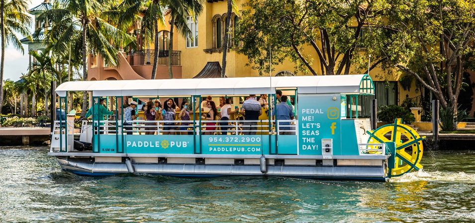 Teal pedal party boat with a rear paddle wheel carrying a group along a sunny tropical canal lined with palm trees and Mediterranean‑style waterfront homes