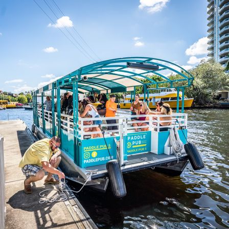 Teal open-air party pontoon tied to a dock as a man secures the line and a group of people board and chat on a sunny urban riverfront.