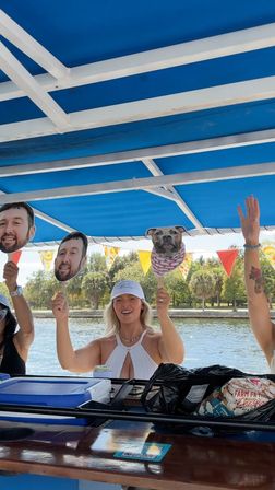 Smiling woman in a white halter top and cap holding novelty face and dog head cutouts on a blue‑canopied party boat, colorful pennant bunting and tree‑lined shoreline in the background.