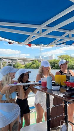 Pontoon boat party on a riverfront: four women in white caps at a bar under a blue canopy, holding shots and colorful drinks with a bridge and city skyline in the background.