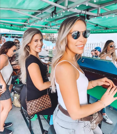 Two smiling women in sunglasses seated at a green‑canopied outdoor pedal‑bar tour, enjoying a casual summer group outing at a counter-style bar bike.