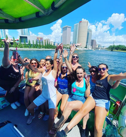 Group of friends cheering on a sunny river boat cruise with the Tampa, Florida downtown skyline, blue sky, and waterfront in the background.