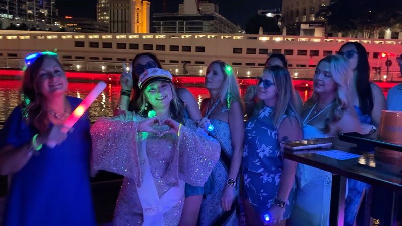 Group of women enjoying a nighttime riverboat waterfront party with city skyline in the background; bachelorette in a captain’s hat and sash makes a heart with her hands while neon and LED lights glow around the group.