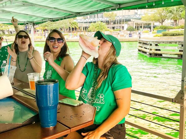 Women in green shirts and beads on a covered waterfront boat bar, one blowing a large conch shell while friends cheer and green-dyed water glows behind them.