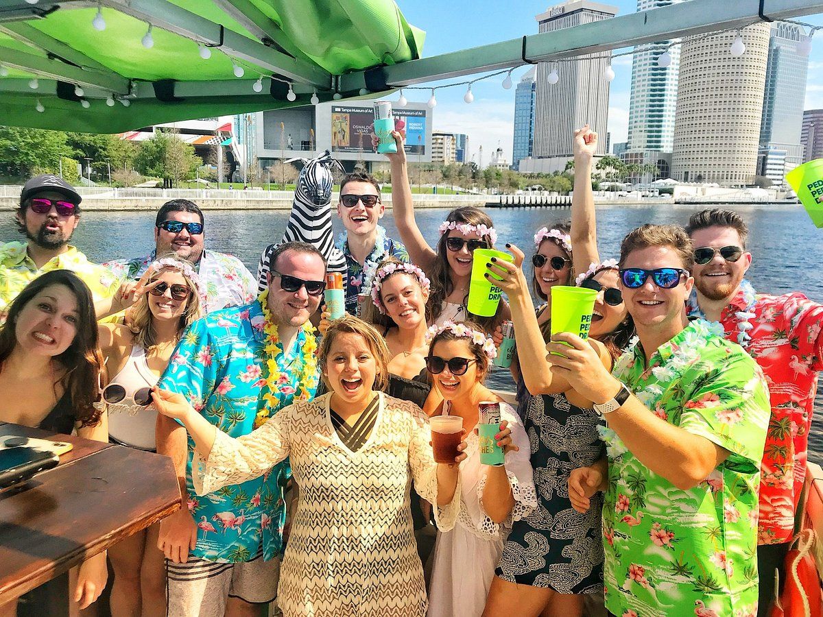 Group of friends in Hawaiian shirts and flower crowns cheering with neon cups on a sunny party boat, inflatable zebra behind them and a city skyline along the waterfront