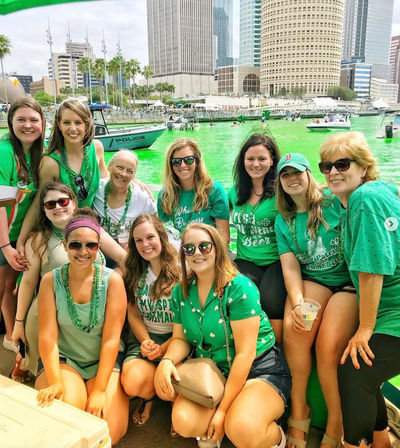 Group of people in green on a boat celebrating St. Patrick’s Day with a bright green-dyed river, police boat, palm trees and downtown Tampa skyline in the background.