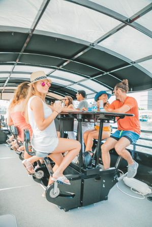 Young adults on a covered pedal-bar boat cruise along a waterfront, pedaling bike-style stools and sipping drinks under a canopy in summer hats and sunglasses