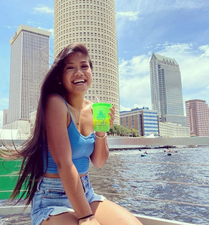 Smiling young woman in a blue crop top and denim shorts holding a bright green cup on a boat with the downtown Tampa skyline and river under a sunny blue sky.