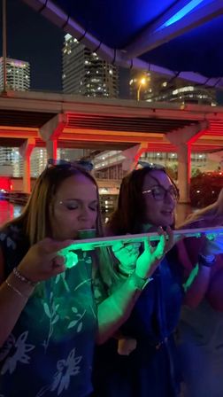 Two people taking a communal shot from an illuminated party plank on a neon-lit urban waterfront at night beneath a highway overpass with high-rise skyline.