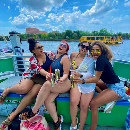 Four friends laughing on a sunny riverboat outing, wearing swimsuits and sunglasses and holding canned drinks with colorful straws, with a tree-lined shoreline, blue sky with puffy clouds and a yellow tour boat in the background.
