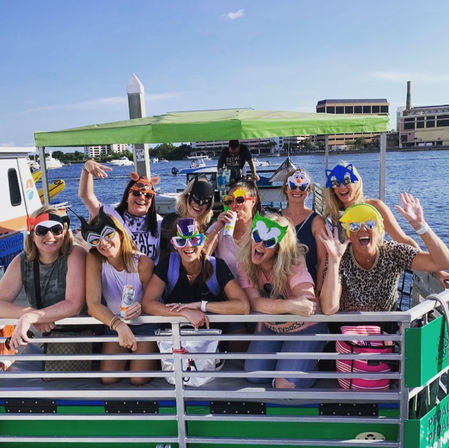 Group of friends wearing colorful novelty masks and sunglasses, laughing and waving on a pontoon boat under a green canopy at a sunny marina with city skyline across the water