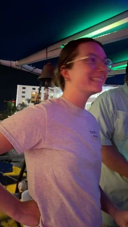 Smiling person in glasses wearing a light gray tee on a rooftop patio under green neon lights at night, with city buildings and outdoor seating visible in the background.