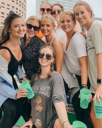 Group of smiling young women on a downtown rooftop posing for a selfie, wearing sunglasses and casual summer clothes while holding bright green drink cups.