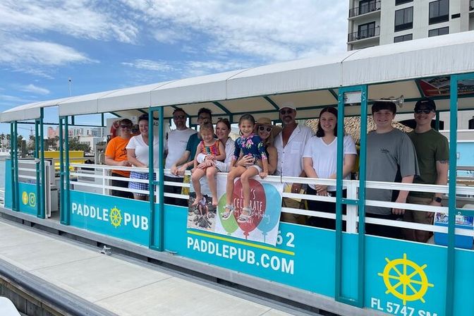 Smiling multi-generational group on a turquoise open-sided tour boat at a sunny marina, two young girls perched on the railing under a white canopy with coastal buildings in the background.