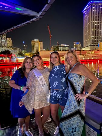 Four women smiling on a nighttime urban waterfront boat cruise with city skyline lights in the background, one wearing a 'Bride to Be' sash.
