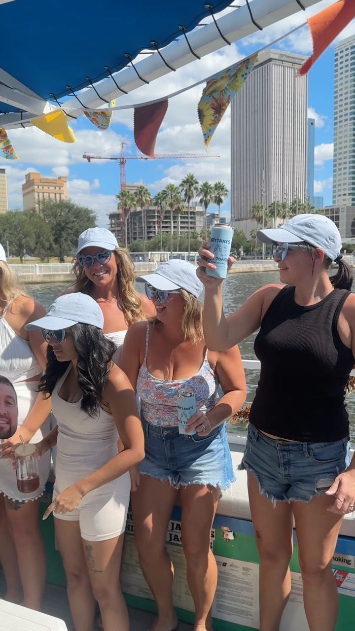 Boat party with a group of women in light-blue caps and sunglasses toasting with canned drinks under colorful pennant flags on a downtown waterfront with palm trees, high-rise buildings, and a crane in the background.