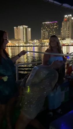 People playing limbo on a boat at night, glittery dress and party lights with a downtown waterfront skyline reflected in the harbor.