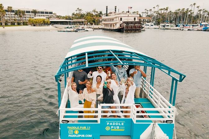 Group of people cheering on a teal-and-white covered pedal-powered party boat cruising a coastal marina, with palm trees, waterfront buildings and docked yachts in the background.