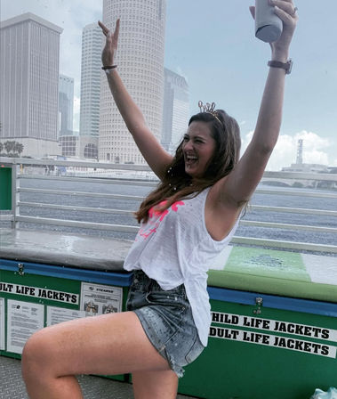 Joyful woman wearing a 'bride' headband and white tank with denim shorts, cheering with arms raised on a waterfront party boat against a city skyline near life-jacket storage.