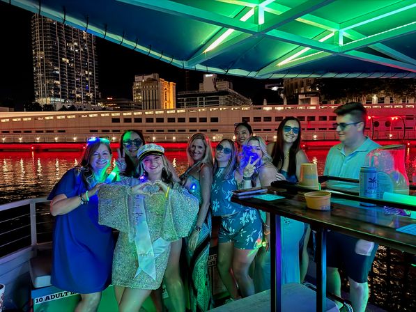 Group of friends posing on a nighttime downtown riverboat cruise under a green neon canopy with city skyline and red-lit waterfront in the background