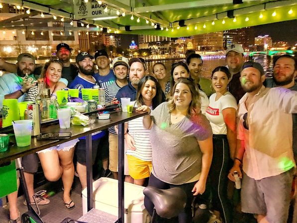 Smiling group of friends on a pedal‑bar boat at waterfront nightlife with city skyline, string lights and drinks
