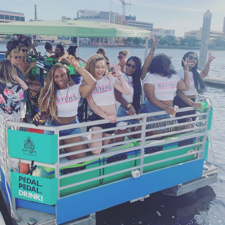 Group of women in matching "Sisters" crop tops partying on a pedal-powered boat at a sunny urban waterfront, laughing and flashing peace signs.