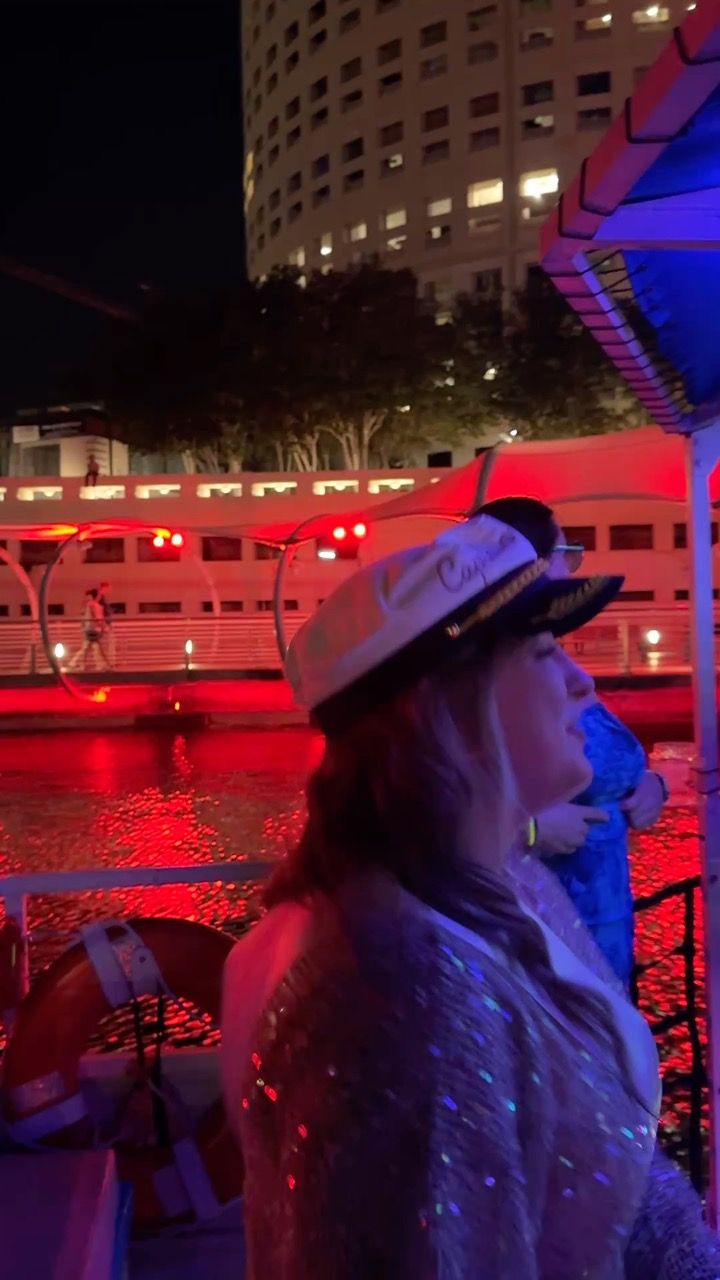 Person in a captain's hat on a nighttime riverboat, red lights reflecting on the water with an illuminated high-rise and riverside walkway in the background.