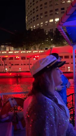 Person in a captain's hat on a nighttime riverboat, red lights reflecting on the water with an illuminated high-rise and riverside walkway in the background.