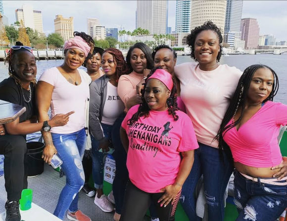 Group of women smiling on a boat with an urban waterfront skyline behind them, several wearing pink shirts—one reads "Birthday Shenanigans Loading"—casual jeans and headwraps for a lively birthday celebration.