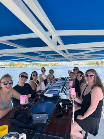 Cheerful group of women on a covered pontoon boat at a waterfront marina, toasting with pink bachelorette cups under a bright blue canopy with city skyline and palm trees in the background.