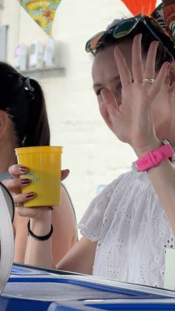 Woman in a white eyelet blouse waving and holding a yellow cup at an outdoor summer party, sunglasses on her head, pink smartwatch and colorful bunting in the background.