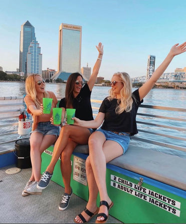 Three women in sunglasses and casual summer outfits laughing and toasting with bright green cups on a boat at sunset, with the Jacksonville riverfront skyline and a steel bridge in the background.
