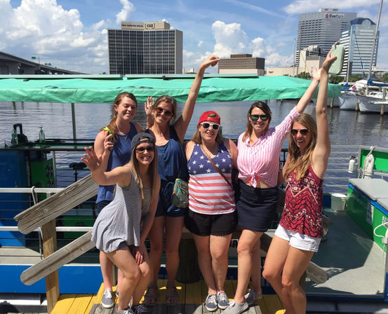 Six friends smiling and waving on a sunny waterfront dock next to a green-canopied boat, with docked sailboats and an urban skyline under blue skies.