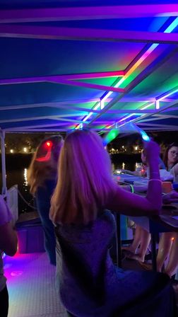Nighttime pontoon boat party with people under a multicolored LED canopy, glow sticks and colorful lights reflecting on the water.