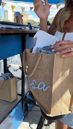 Sunny waterfront scene: hands reach into a jute beach tote containing a patterned towel and boxed items at a dockside bar, with bunting and marina buildings in the background.