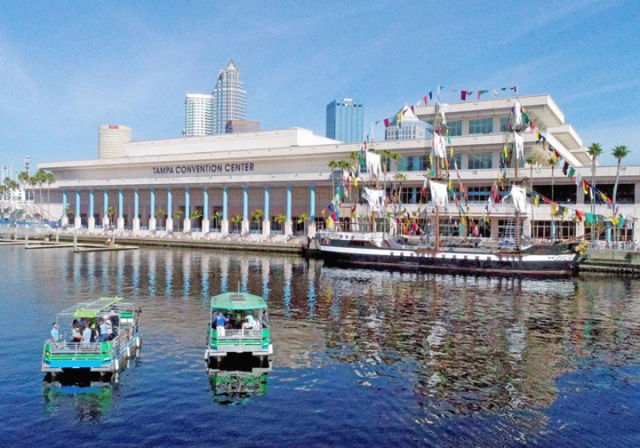 Tampa waterfront scene with a riverside convention center, a tall ship draped in colorful flags, two green tour boats and downtown skyline reflected in the harbor