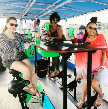 Three friends smiling and holding neon green cups aboard a canopy-covered, pedal-powered party bar with bike seats and a central wooden tabletop during an outdoor daytime outing.