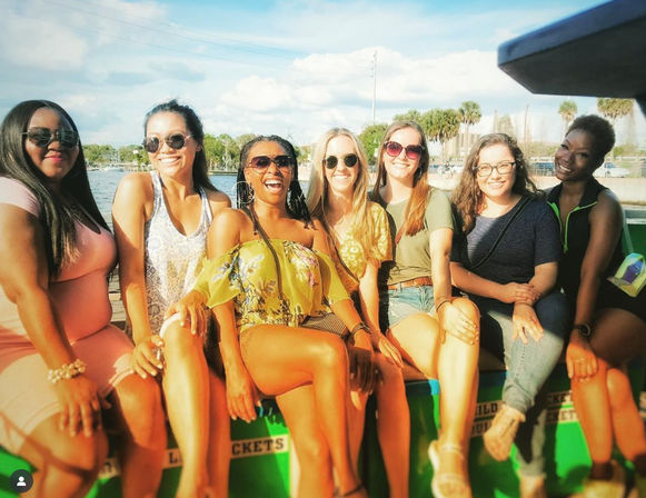 Seven smiling friends relaxed on a colorful boat at a sunny waterfront lined with palm trees, wearing sunglasses and summer outfits