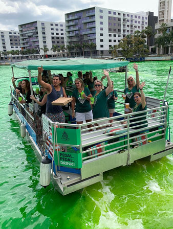Pontoon boat party with a group of adults waving and holding drinks on an urban waterfront dyed bright green for St. Patrick's Day.